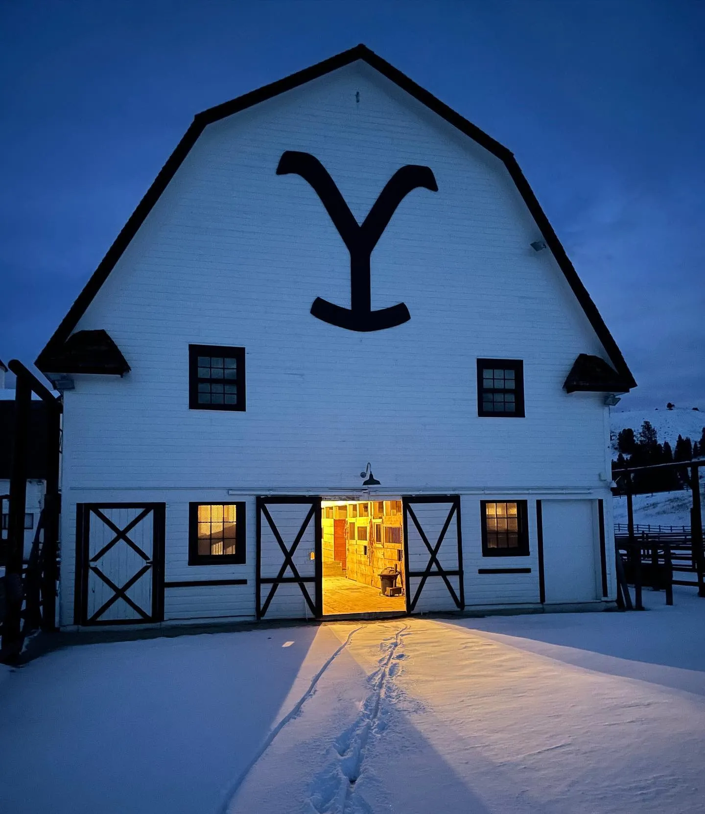 The white barn at dusk in snow, its doors lit and a large black Y brand under the roof peak