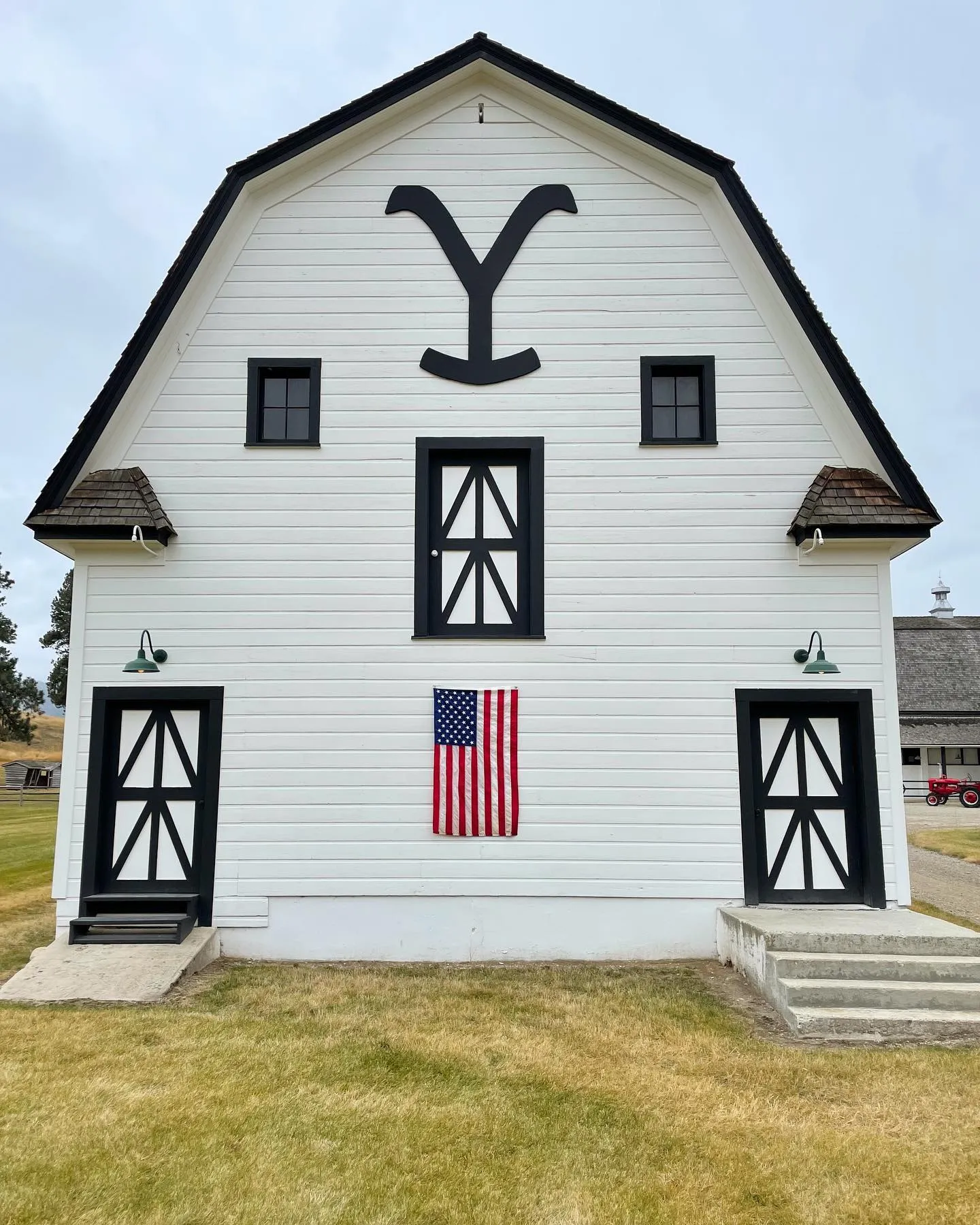 The white gambrel end of a ranch barn today, with black trimmed windows and doors, a large black Y brand under the roof peak and an American flag between the doors