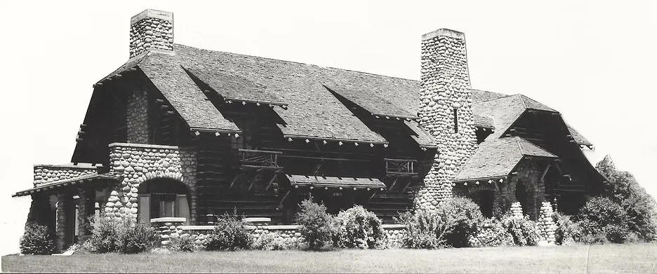 Black and white archival photograph of the Ford-Hollister Lodge: a long log building with deep shingled roofs, two tall river stone chimneys and stone arched porches, framed by shrubs