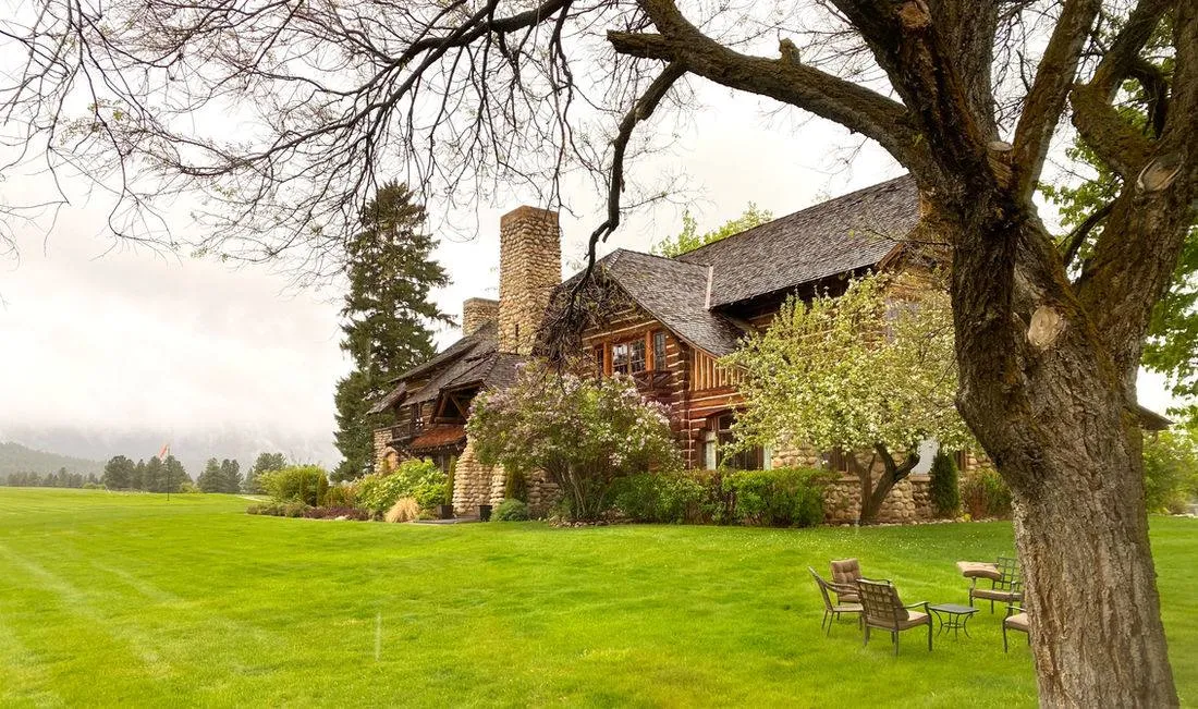 The log and stone lodge beside a large shade tree, across a green lawn