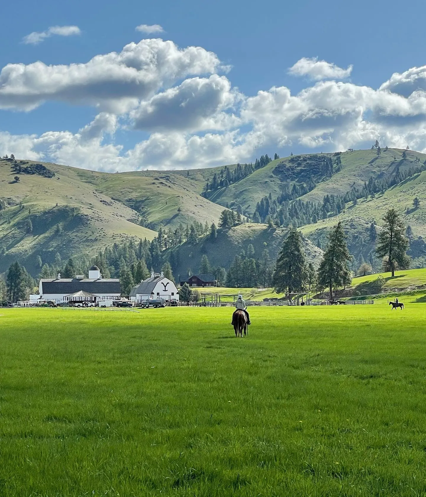 A rider crossing the green meadow toward the white barns, with the hills of the Bitterroot Valley behind