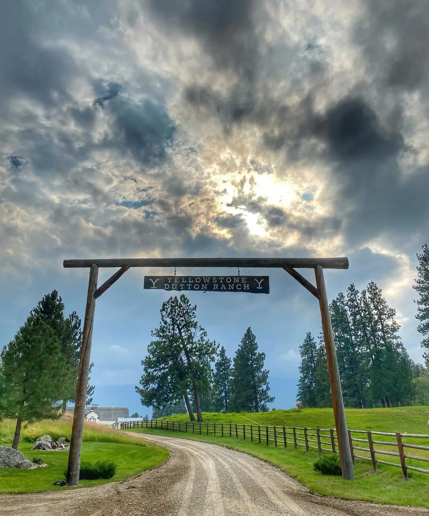 A tall timber gate over a gravel ranch road under a dramatic cloudy sky, its hanging sign reading Yellowstone Dutton Ranch, with pines and a split rail fence