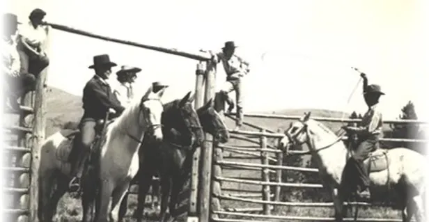 Sepia archival photograph of riders in cowboy hats on light and dark horses at a log corral, one rider swinging a rope while others sit on the top rail